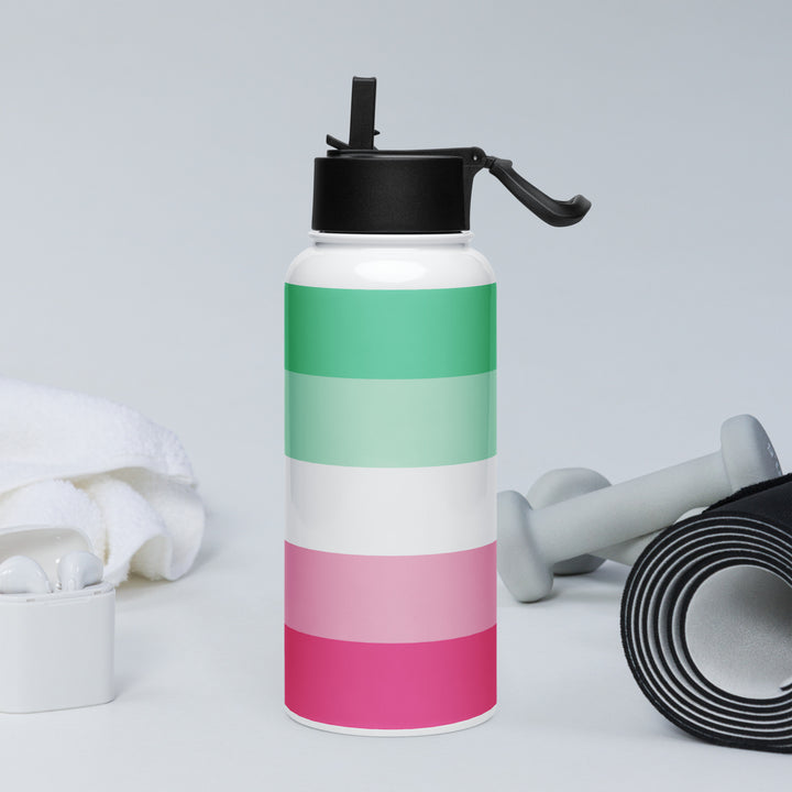 Stainless steel water bottle with a XXXXXX Pride Flag printed around it, on a rock with a background of fitness equipment. By The Rainbow Stores