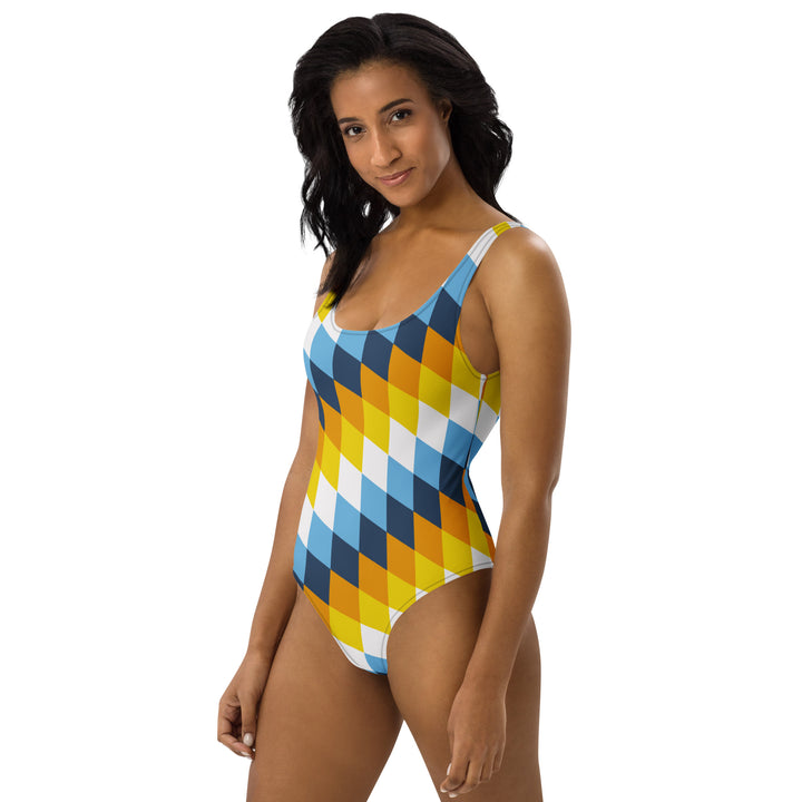 A woman or non binary person with long dark hair is wearing a one-piece swimsuit with a diamond pattern angled top left to bottom right. The interlocking diamonds are in layered colours, and the layers are made from the colours of the Aroace  Pride Flag 
