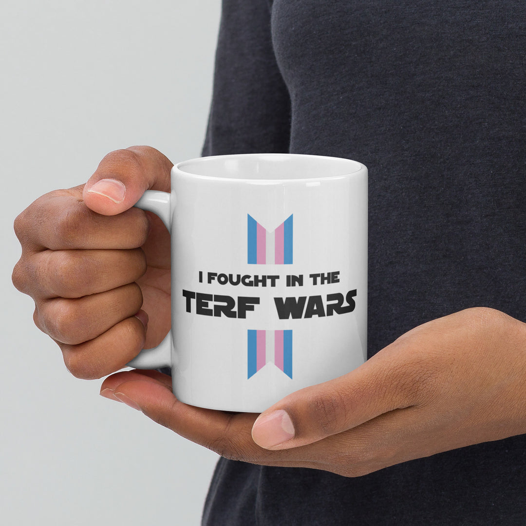 A mans hands holding a white mug with the words I Fought In The TERF Wars with a trans flag ribbon top and bottom of the text. by The Rainbow Stores
