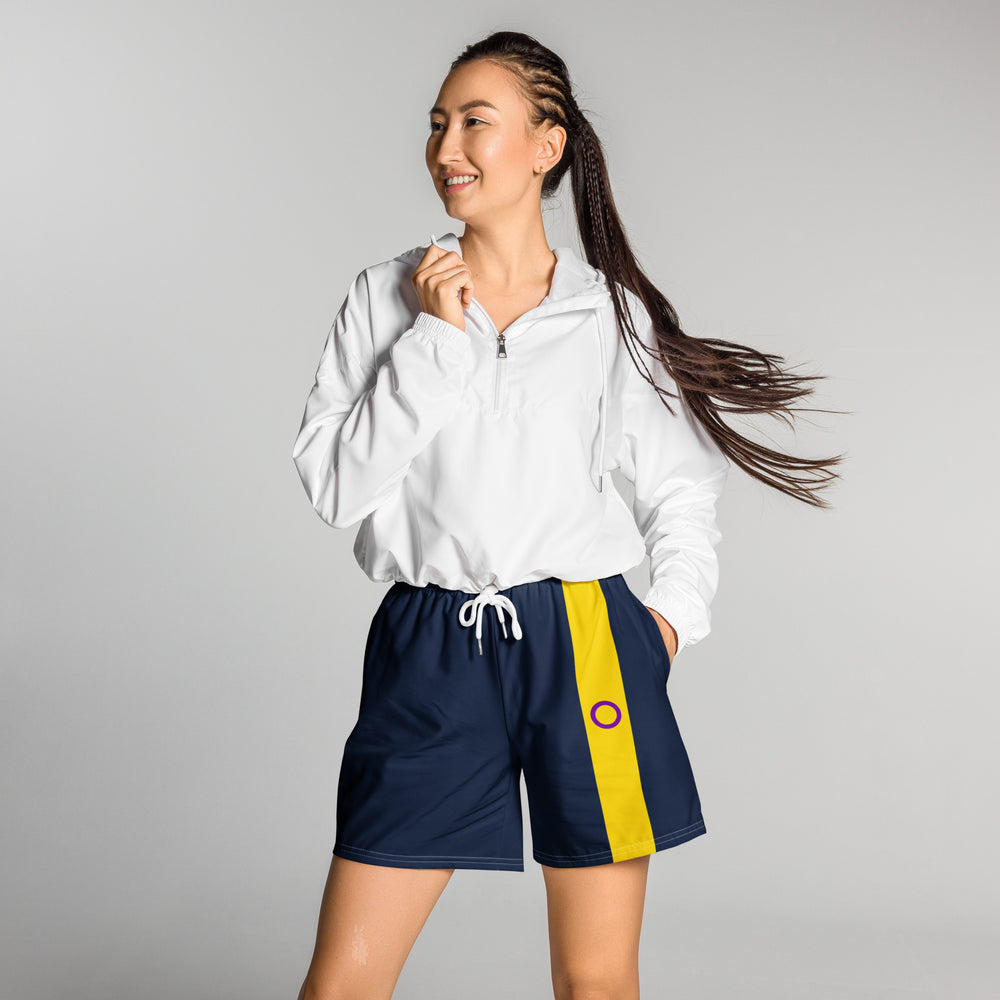 A person with long brown braided hair stands against a grey background and looks to the left of the image. She/they have their right hand holding the collar of a short white zipped hoodie, and their left hand in the pocket of a pair of dark blue long shorts. On the left leg of the shorts is a vertical stripe in the colours of the Intersex pride flag running left to right.