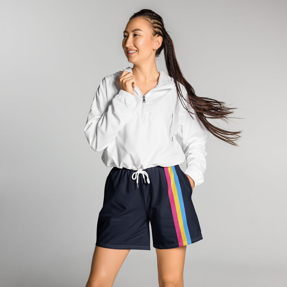 A person with long brown braided hair stands against a grey background and looks to the left of the image. She/they have their right hand holding the collar of a short white zipped hoodie, and their left hand in the pocket of a pair of dark blue long shorts. On the left leg of the shorts is a vertical stripe in the colours of the Pansexual pride flag running left to right.
