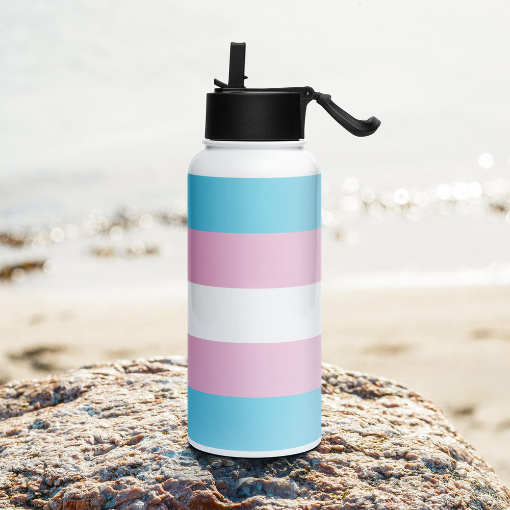 Stainless steel water bottle with a Trans Pride Flag printed around it, on a rock with a beach background. By The Rainbow Stores