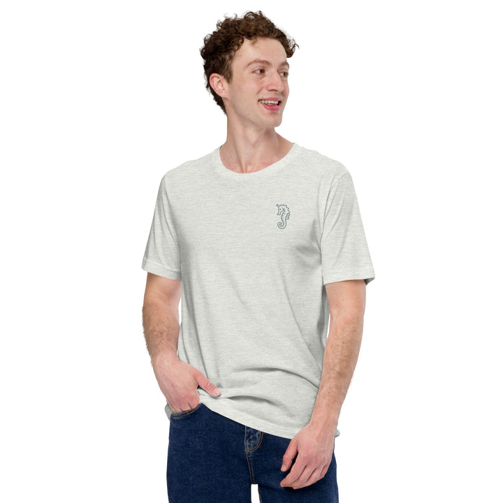 A smiling man with curly brown hair and jeans wears a light grey t-shirt with a small mid grey trans dad seahorse motif on the left chest