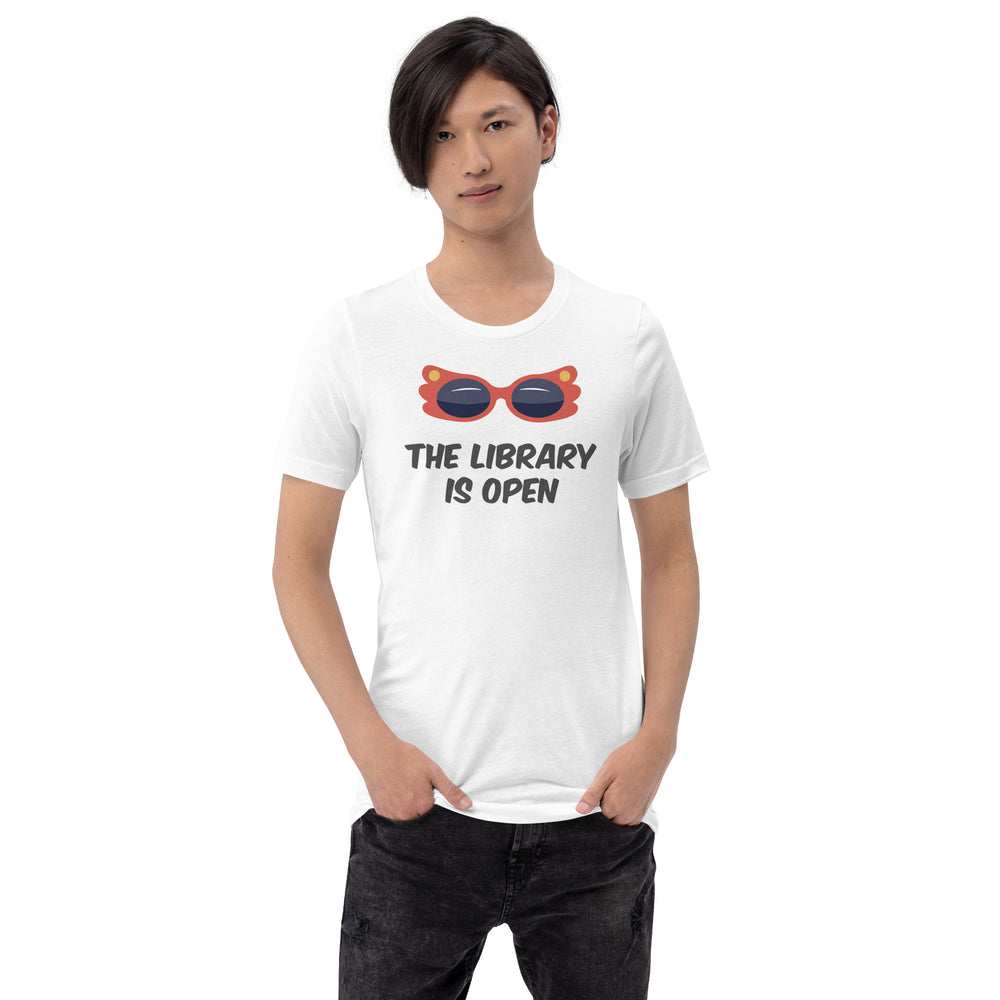 A man wearing a white t-shirt with an illustration of a pair of camp red sunglasses on, and underneath are the words The Library Is Open in black. By The Rainbow Stores
