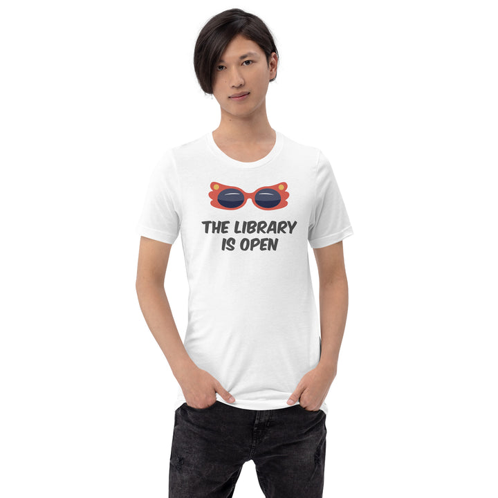 A man wearing a white t-shirt with an illustration of a pair of camp red sunglasses on, and underneath are the words The Library Is Open in black. By The Rainbow Stores